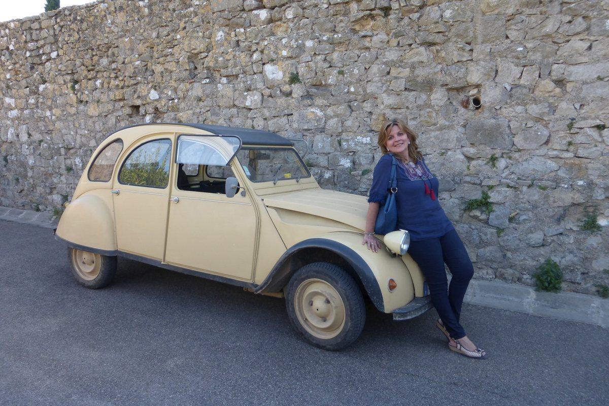 A French learner smiles as she leans on a beige Citroën 2CV in front of a stone wall during an excursion in the Aude